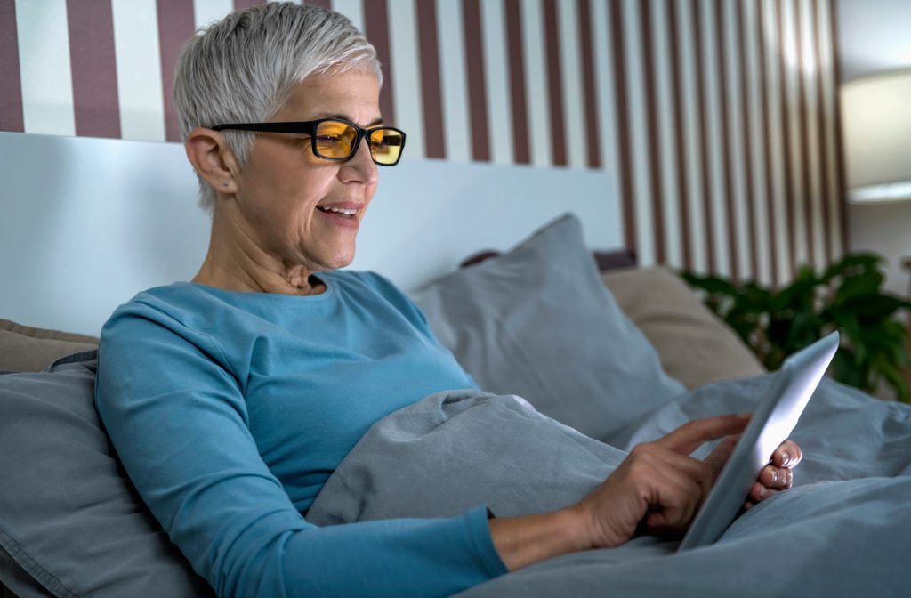 A senior wearing blue light blocking glasses while in relaxing in bed and browsing on her iPad.