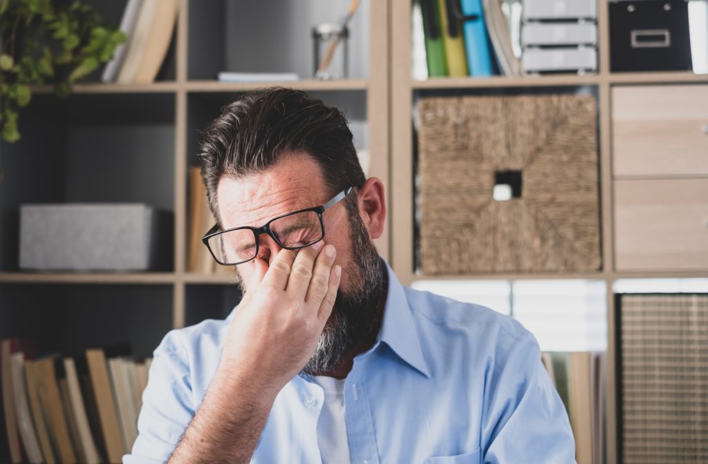 Stressed man rubbing eyelids under his glasses, suffering from dry eyes syndrome due to long computer overwork, massaging nose bridge relieving pain at office at home.