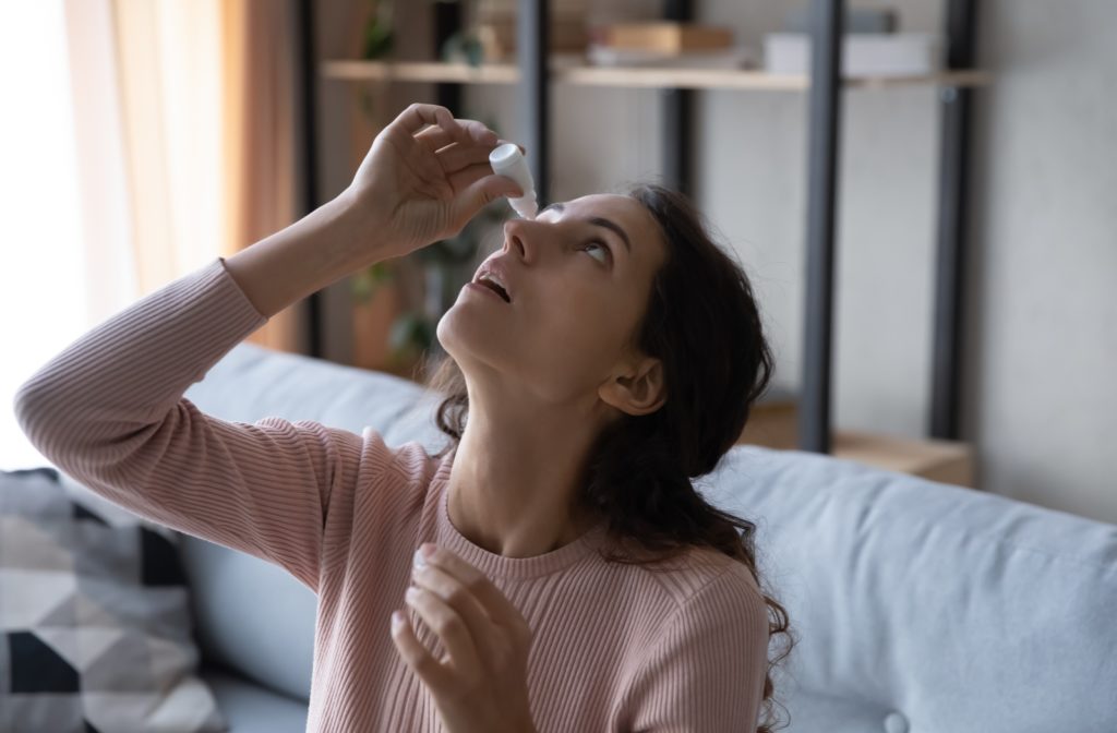 Young woman sitting at home on her couch putting eye drops into her right eye to help with irritation.