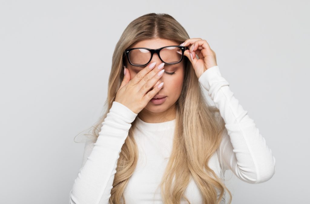 A woman with a white shirt in front of a grey background holding her right eye in discomfort as her eyes are adjusting to her new glasses