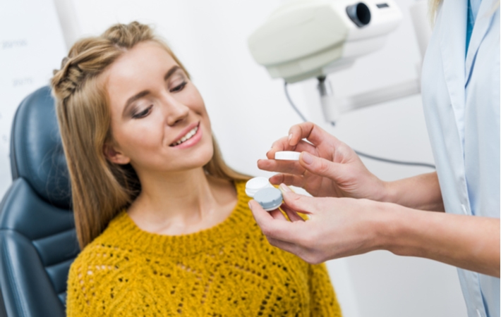 A woman with a yellow shirt on sitting in a chair talking to her optometrist while they hold a container of contact lenses after her contact lens exam