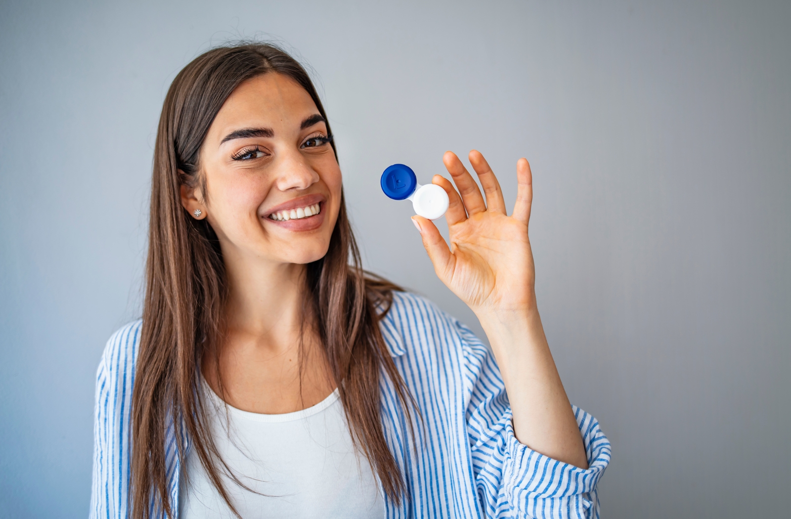 A woman in a blue and white shirt holding up in her left hand a blue and white contact lens container while smiling