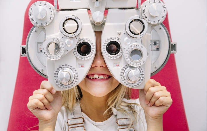 A young girl smiling while behind a phoropter as she is visiting her optometrist for her regular eye exam