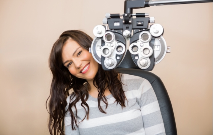 A woman seated behind a phoropter to measure her eye prescription at her regular eye exam