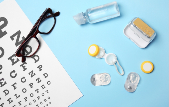 A set of contact lenses with their case and solution, positioned beside a pair of glasses that are resting on top of a Snellen eye chart