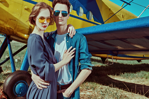 A man and a woman wearing aviator sunglasses posing in front of an older style plane