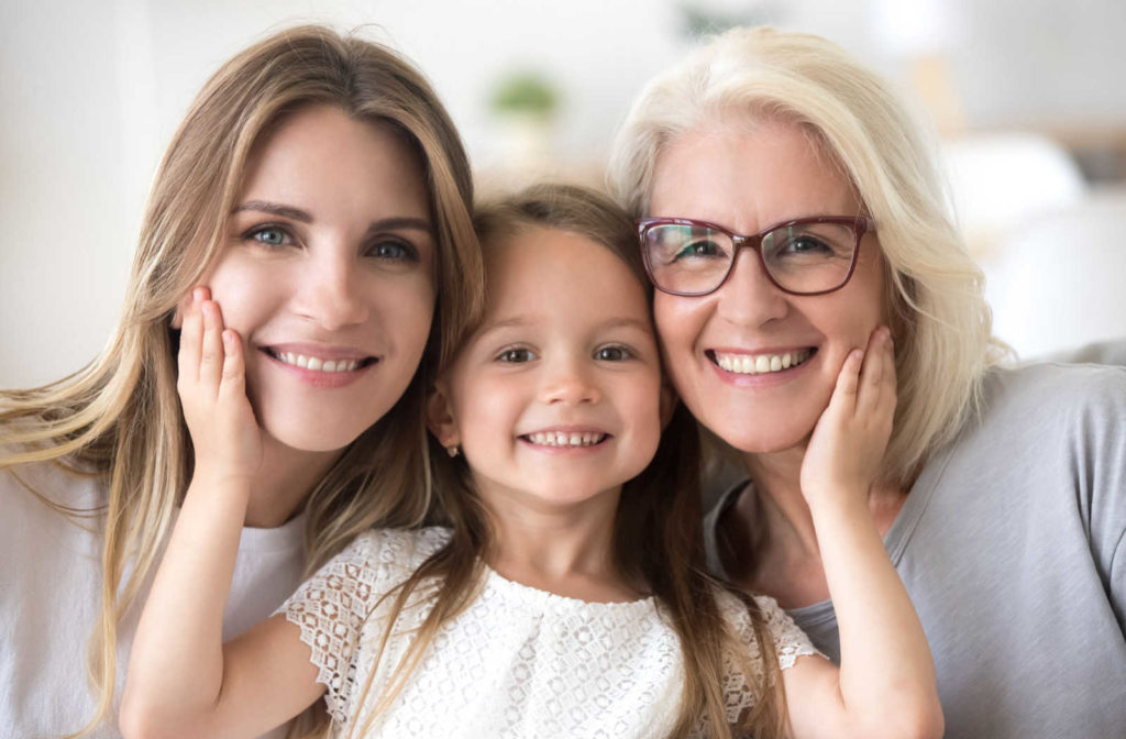 A grandmother wearing glasses, a mother, and a young daughter in the middle with her hands on their cheeks all smiling.