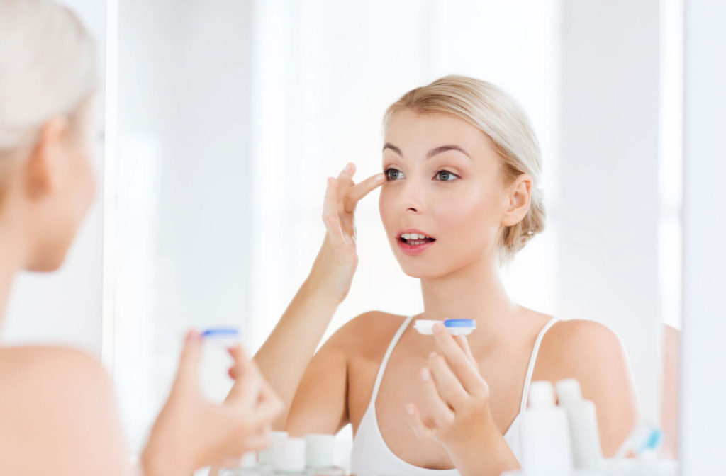 A woman looking in the mirror and inserting a contact lens in her left eye using her left middle finger. She is holding a contact lens case in her right hand.