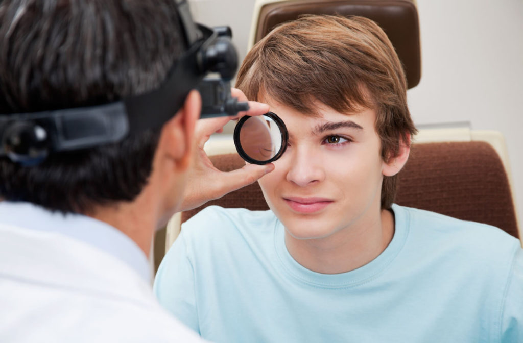 A male eye doctor is holding a magnifying glass and performing a dilated eye exam on a male patient in a light blue coloured shirt.