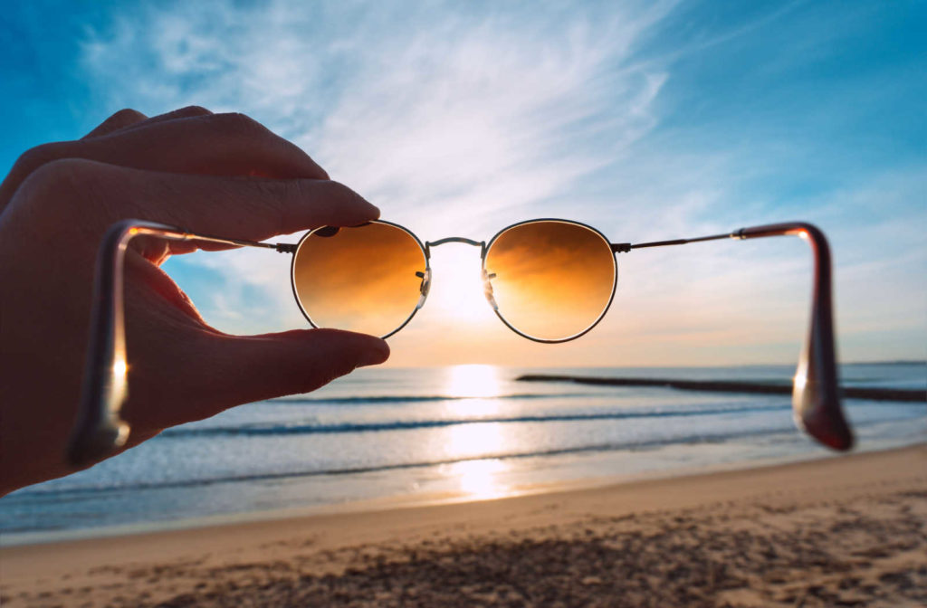 A hand holding up a pair of sunglasses to the sun while on the beach to display how sunglasses block harmful UV rays from eyes