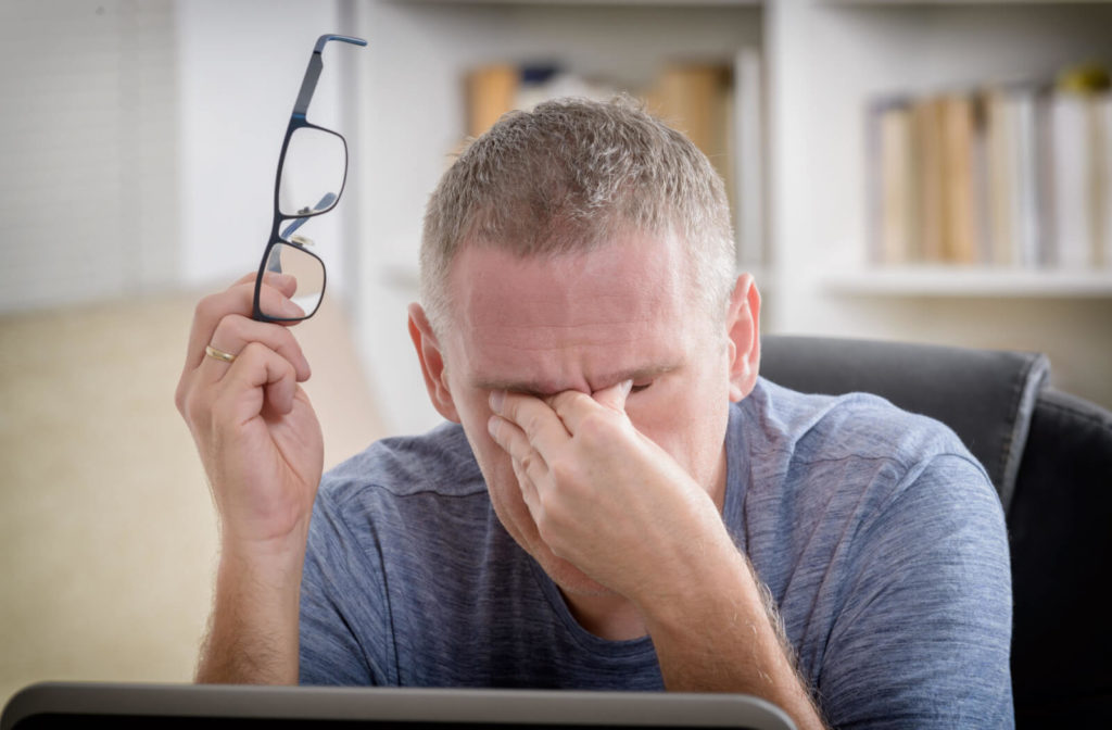 A man removing his glasses to rub his eyes after prolonged use of a laptop screen.