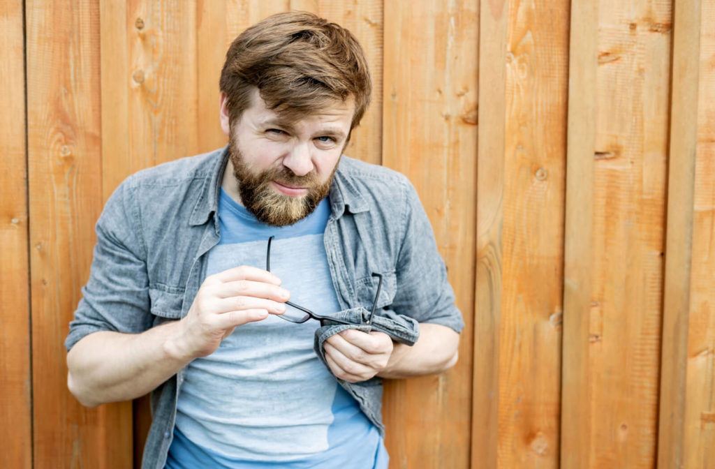 A bearded man leaning against a wooden fence cleans his glasses with his shirt.