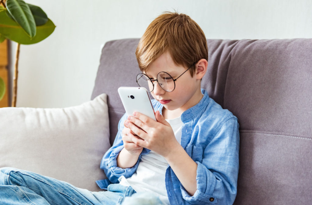 A young boy wearing glasses sits on a couch, using a smartphone too close to his face.
