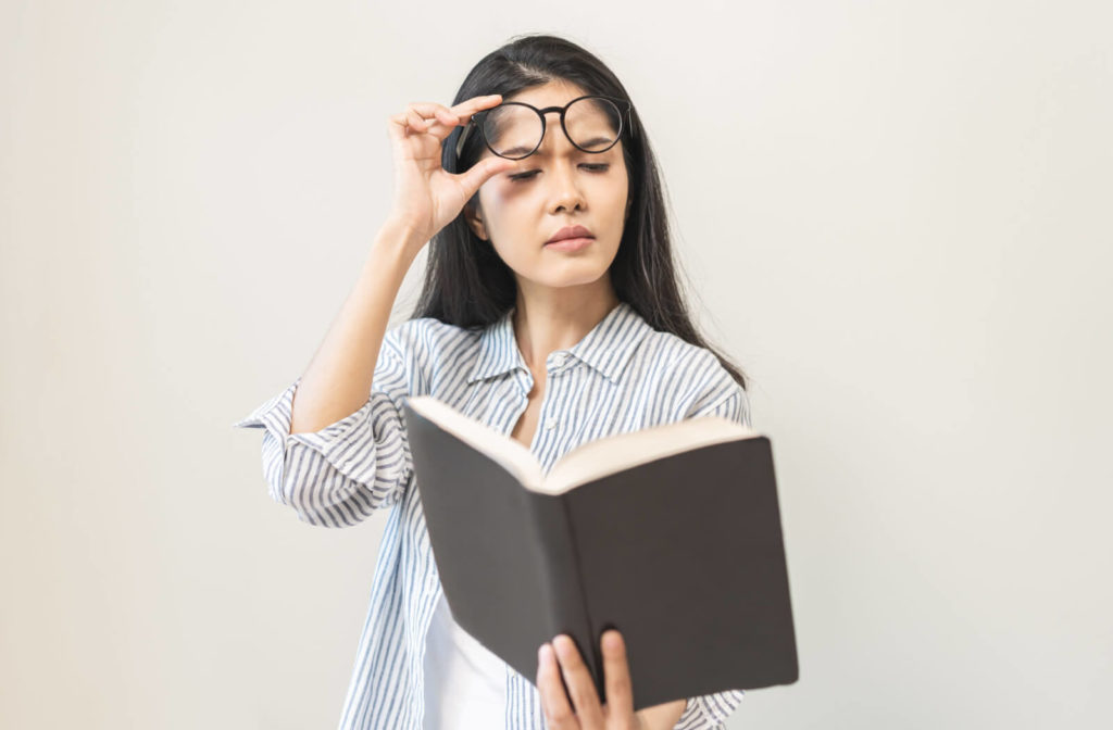 A mature woman takes off her eyeglasses and holds a book at a distance while reading it.