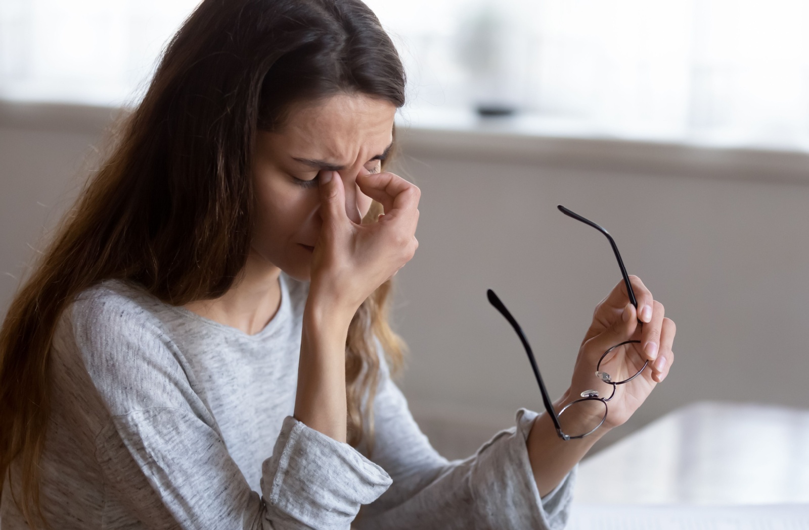 Young woman rubbing here eyes from dry eyes, holding her glasses in one hand while working on laptop.
