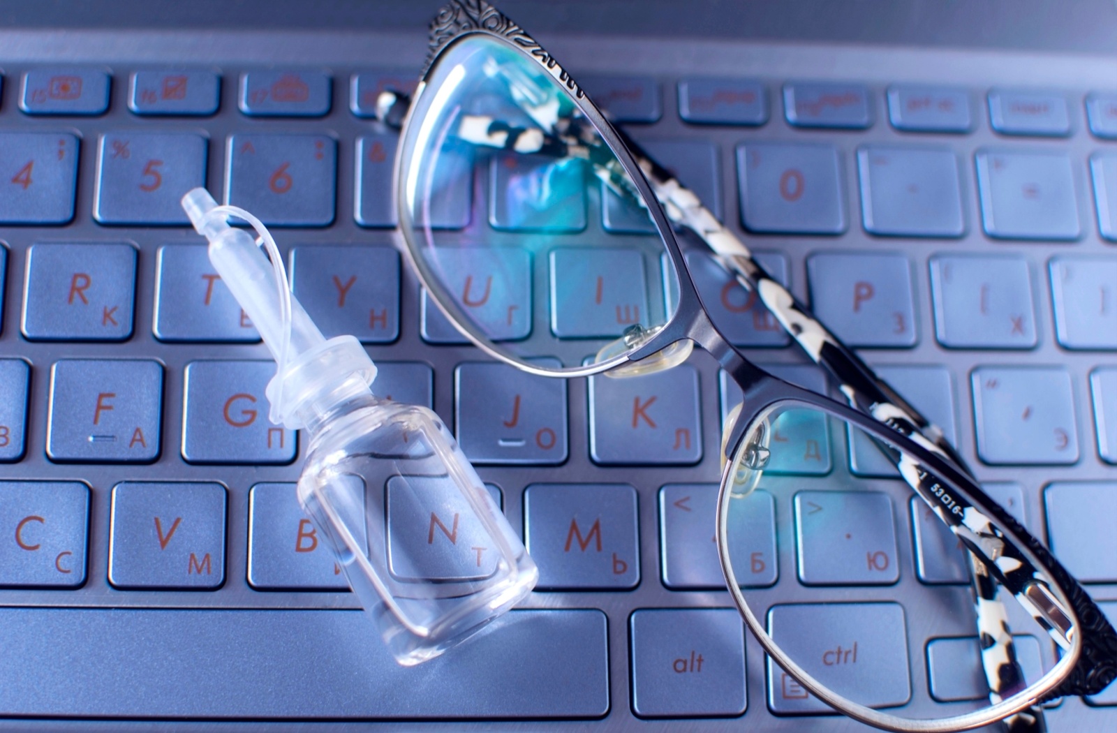 Close-up of a pair of glasses and a bottle of eye drops resting on a laptop keyboard in blue light