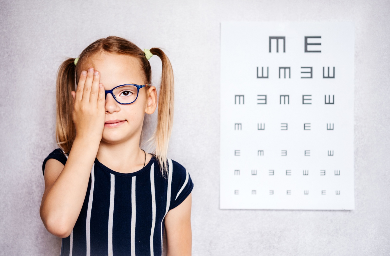 A child covers one eye while standing in front of an eye chart.