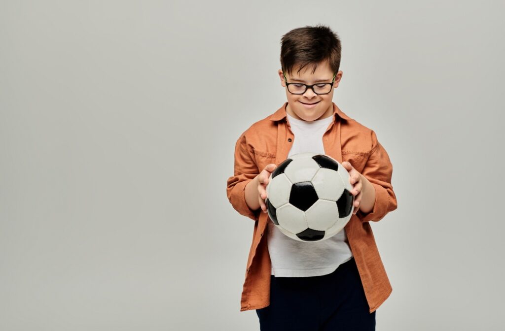A child wearing glasses holding a soccer ball while looking down at it, standing against a plain background.